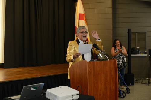 Emcee gesturing animatedly at the podium with trophies displayed