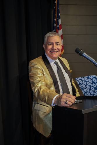 Host in a gold jacket poses at the podium with the flag